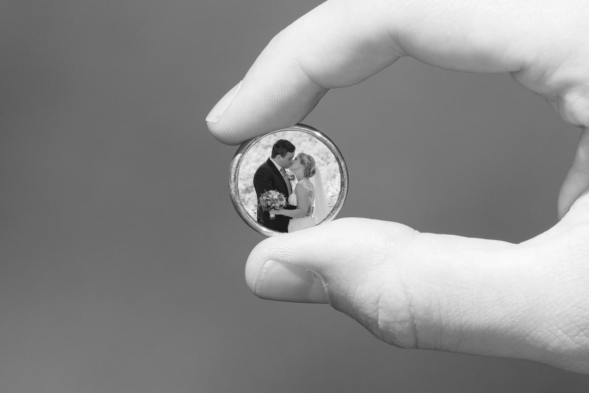 hochzeitsbilder - sichtbare momente - küssendes brautpaar bild montiert in einen ring, der zwischen daumen und zeigefinger gehalten wird
